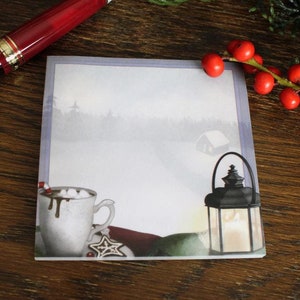 Peut inclure: Un bloc-notes sur le thème de l'hiver avec un paysage enneigé, une tasse de chocolat chaud avec des guimauves et une lanterne. Le bloc-notes est sur une surface en bois avec une branche de baies rouges.