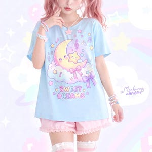 Könnte beinhalten: Hellblaues T-Shirt mit einem Cartoon-Mond- und Stern-Design, Wolken, Noten und den Worten "Sweet Dreams". Das Shirt wird mit rosa Shorts mit Rüschensaum getragen.