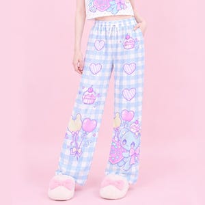 May include: Light blue and white gingham pyjama trousers featuring a drawstring waist and a repeating pattern of pink hearts, cupcakes, and a blue bear character holding balloons. The trousers have a wide-leg silhouette and are paired with fuzzy slippers.