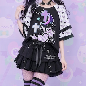 May include: A black and white raglan t-shirt featuring a graphic of a crescent moon, crosses, and a winged heart, paired with a black tiered mini skirt with a lace-up front, silver metal rings, and the text Merbunny BABY embroidered in white.
