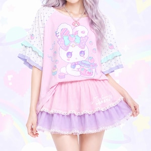 May include: A light pink t-shirt with a graphic of a white bunny holding a jar of strawberries, featuring polka-dot sleeves with ruffled cuffs. The outfit includes a layered pink and purple tulle skirt with white lace trim and the text Merry Baby.