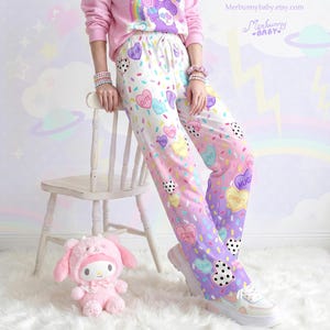 Può includere: Una persona indossa una felpa rosa e pantaloni del pigiama. I pantaloni del pigiama hanno una base bianca con motivi colorati a cuore e pois. I pantaloni passano dal bianco al rosa e al viola. Un peluche rosa è sul pavimento.