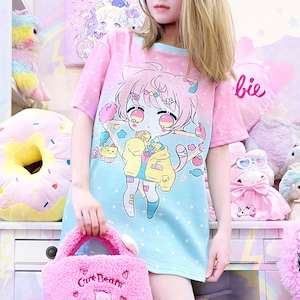 Può includere: Un abito a maglietta rosa e blu con una grafica di gatto dei cartoni animati, abbinato a una borsa tote rosa e soffice. La borsa tote presenta il testo "Care Bears" e un design ricamato di Care Bear. Lo sfondo include peluche e una mensola decorativa.