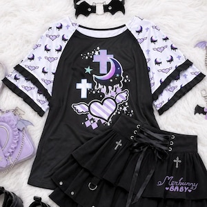 May include: A black and white raglan t-shirt with a purple crescent moon, crosses, and winged heart graphic. A black tiered skirt features a lace-up front, silver cross charms, and the text Merbunny BABY. Includes a black bat-wing choker.