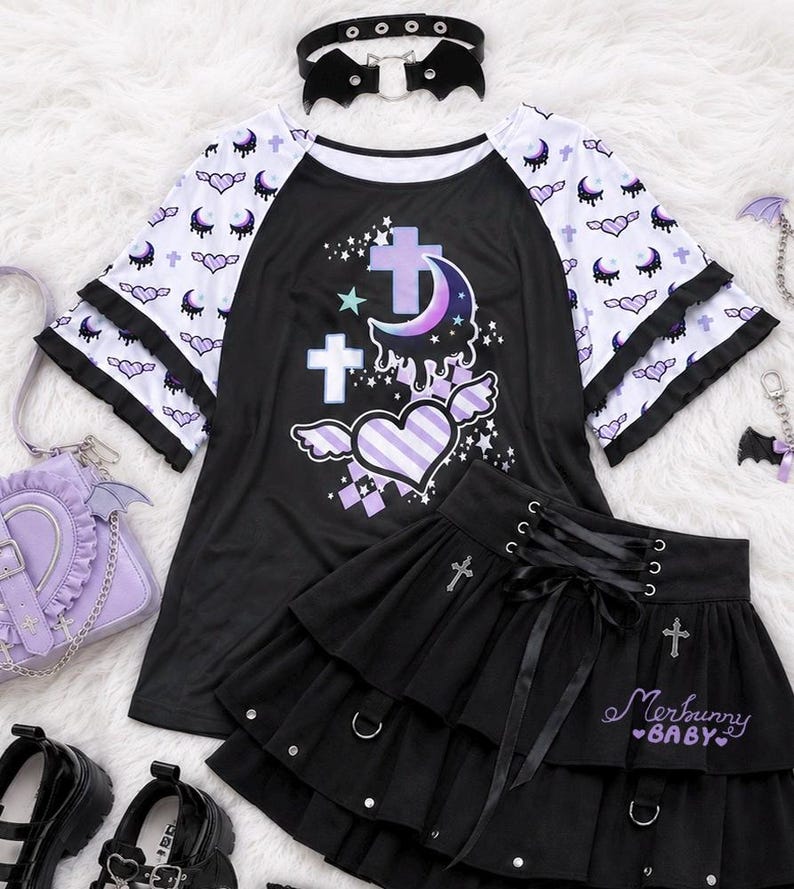 May include: A black and white raglan t-shirt with a purple crescent moon, crosses, and winged heart graphic. A black tiered skirt features a lace-up front, silver cross charms, and the text Merbunny BABY. Includes a black bat-wing choker.