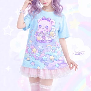 May include: Light blue t-shirt dress with a cartoon bear graphic. The bear is in a basket with stars and rainbows. The dress has a pink lace trim and the text "Merbunny BABY".