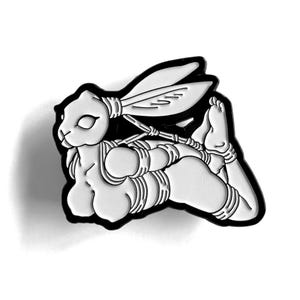 Könnte beinhalten: Ein schwarz-weißer Emaille-Pin mit einer stilisierten Illustration eines Kaninchens in einer Bondage-Pose. Das Kaninchen ist weiß mit schwarzen Umrissen und Details und mit Seilen gefesselt. Der Pin hat einen schwarzen Rand.