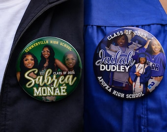 May include: Two circular pin-back buttons featuring high school graduation photos. The left button is green with the text Summerville High School, Class of 2025, and Sabrea Monaé. The right button is blue with Class of 2023, Jailah Dudley, and Apopka High School.