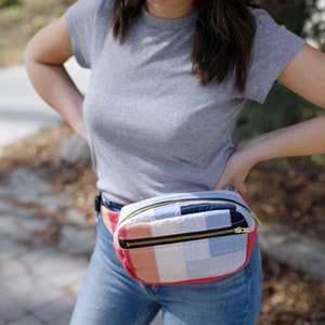 May include: A quilted fanny pack with a zipper closure. The pack is made of patchwork fabric in shades of blue, pink, and white. The pack is worn around the waist of a person wearing a grey t-shirt and blue jeans.