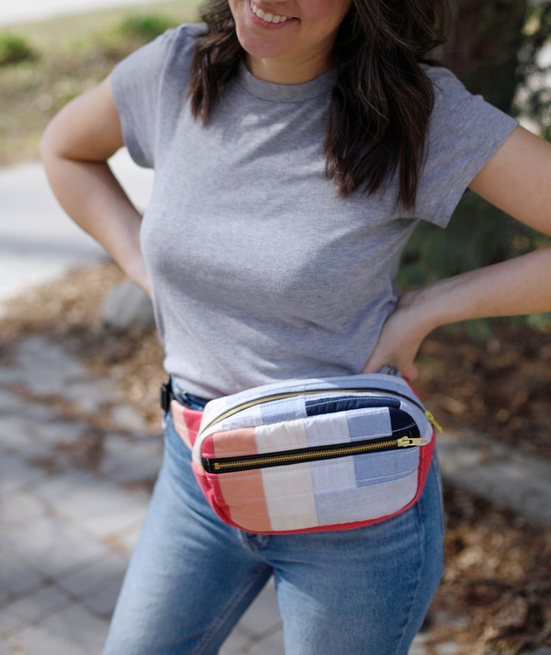 May include: A quilted fanny pack with a zipper closure. The pack is made of patchwork fabric in shades of blue, pink, and white. The pack is worn around the waist of a person wearing a grey t-shirt and blue jeans.