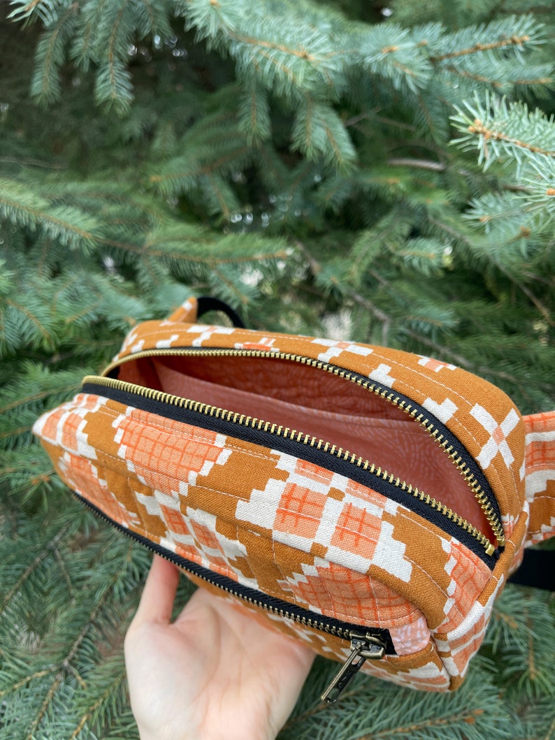 May include: A brown and orange patterned fabric fanny pack with a black zipper and gold zipper pull. The fanny pack is open and the inside is lined with a light pink fabric.
