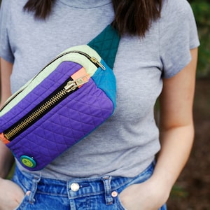 May include: A purple, green, and blue quilted fanny pack with a gold zipper and a green smiley face button. The fanny pack is worn around the waist of a person wearing a grey t-shirt and blue jeans.