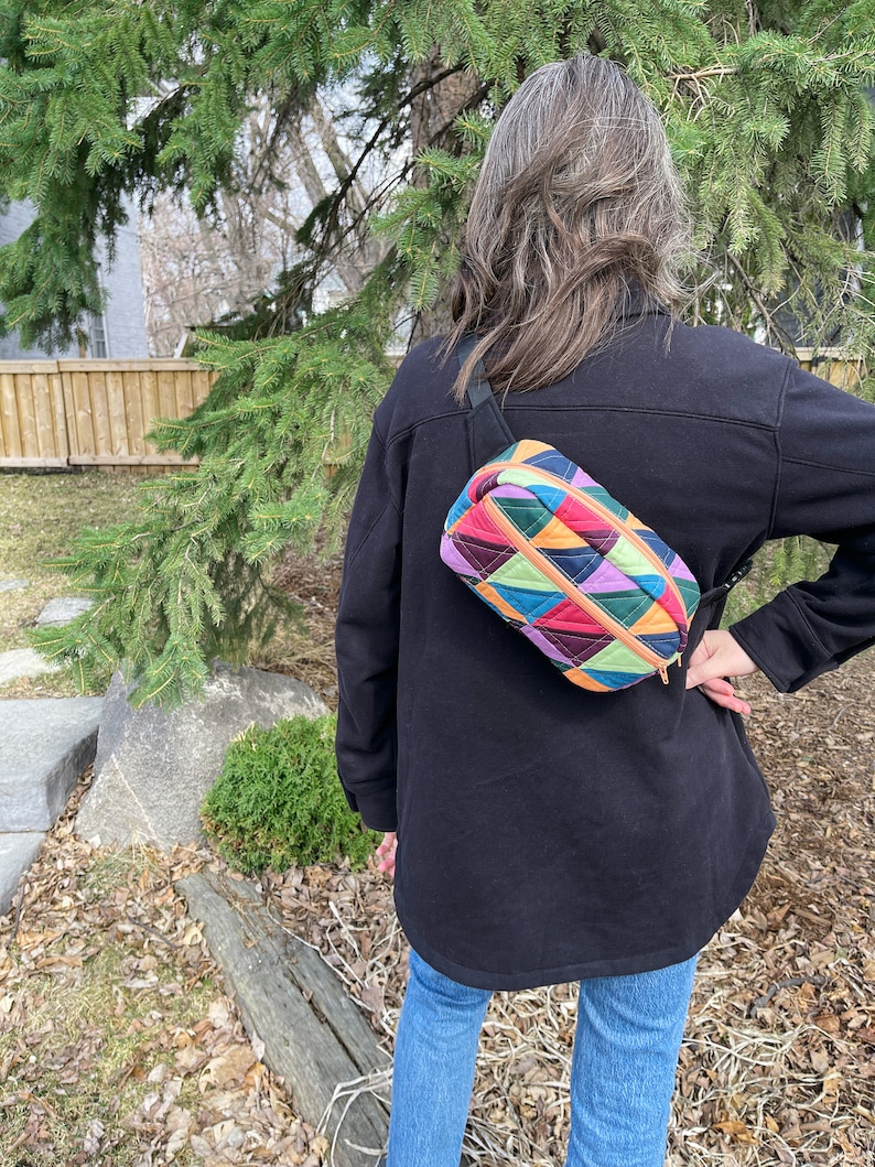 May include: A colorful quilted fanny pack with a geometric pattern in shades of green, red, orange, purple, and blue. The fanny pack is worn across the body of a person wearing a black jacket and blue jeans.