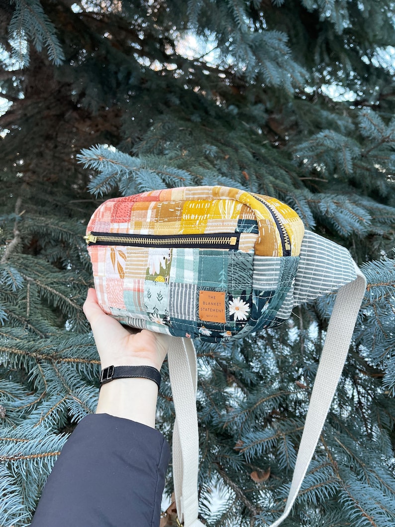 May include: A patchwork fabric fanny pack with a zipper closure and a white adjustable strap. The fanny pack has a brown leather patch with the text "The Blanket Statement" on it.