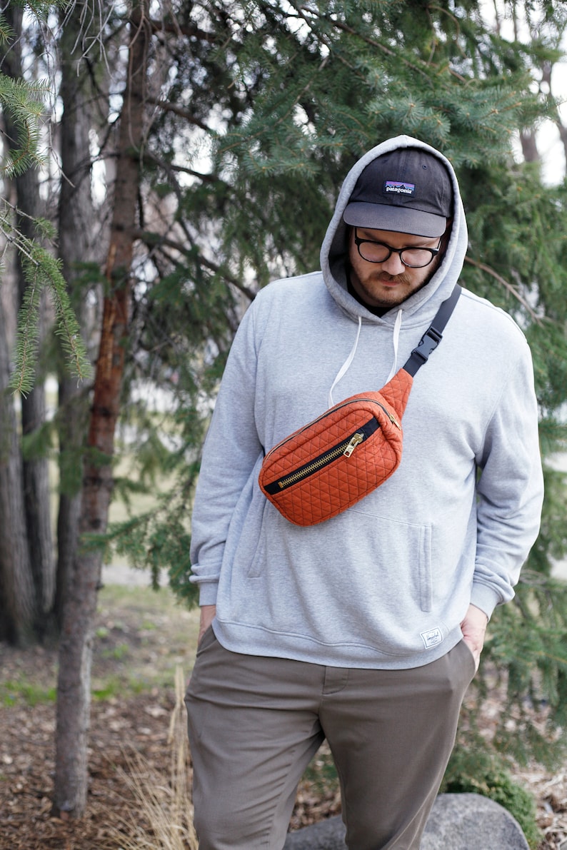 May include: A person wearing a gray hoodie with a black hat and a bright orange fanny pack. The fanny pack has a zipper and a black strap.