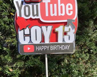 Puede incluir: Un adorno para pastel de YouTube de color rojo y blanco con un pulgar hacia arriba, una campana y un icono de flecha. El adorno dice "You Tube Coy 13 Happy Birthday".