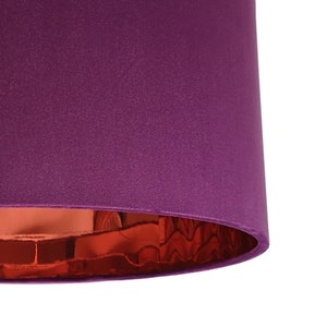 May include: A close-up of a cylindrical lampshade. The exterior is a deep purple, with a textured surface. The interior is a shiny, reflective copper colour, creating a contrast. The lampshade is against a white background.