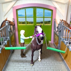 Può includere: Una scena di scuderia giocattolo con un cavallo marrone e bianco con una cavezza viola. Una figura tiene le redini del cavallo. La stalla ha porte e finestre verdi, con altri cavalli giocattolo nelle stalle. La scena è ambientata su un pavimento di mattoni grigi.