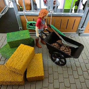 May include: A farm playset featuring a miniature figure with a black wheelbarrow filled with brown objects. Yellow and green hay bales are stacked on a brick surface. A barn with stalls is in the background.