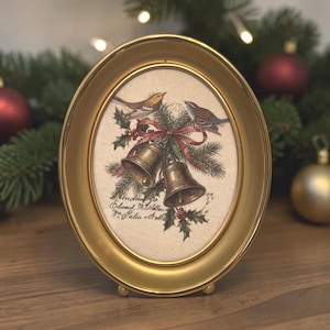 May include: An oval gold-toned frame containing a print of two brass bells tied with a red ribbon, surrounded by pine branches, holly berries, and two small birds. The print includes handwritten-style text at the bottom and top right.