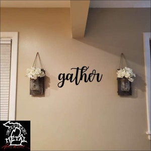 May include: A black metal sign with the word "gather" in cursive script, hanging on a beige wall with two mason jars filled with white flowers hanging on either side of the sign.