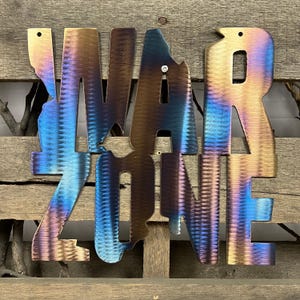 May include: Metal wall art with the words "WAR ZONE" in a blue, purple, and brown color scheme. The letters are cut out and have a textured finish.