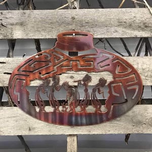 May include: A metal wall art piece depicting a Native American dance scene. The art is oval shaped and features a rusty brown color with a layered, textured finish.