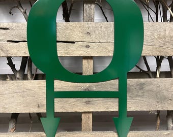 Puede incluir: Corte de metal verde de la letra "O" con dos flechas apuntando hacia abajo unidas a la parte inferior. La letra está en un estilo de fuente redondeado.