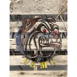 May include: A metal wall art piece depicting a bulldog with red eyes and a barbed wire collar. The bulldog is facing the right side of the image. The words "USMC" are displayed in gold letters below the bulldog.