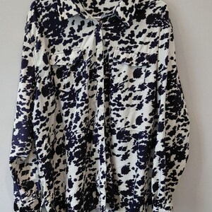 May include: A long-sleeved button-down shirt featuring a black and white cow print pattern. The shirt includes a standard collar, a front button placket, and two chest pockets with buttoned flaps. The interior label reads Crazy Train.