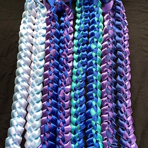 May include: Four braided satin ribbon leis in shades of blue and purple. Each lei is made of interwoven ribbons, with a loop at the top for hanging. The colors include light blue, royal blue, teal, and various shades of purple.
