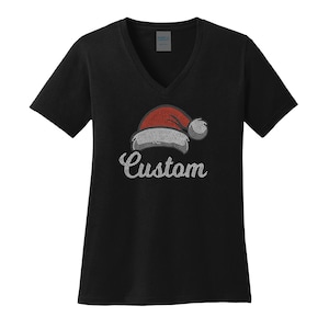 Puede incluir: Camiseta negra con cuello en V con un diseño de gorro de Papá Noel. El gorro es rojo y blanco con un pompón blanco. La palabra "Custom" está escrita debajo del gorro en cursiva.