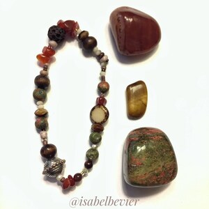 May include: A bracelet made with various stones and beads, including red carnelian, green aventurine, tiger's eye, and lava rock. The bracelet is displayed with three tumbled stones: red carnelian, tiger's eye, and green aventurine. The image includes the text "@isabelbevier".