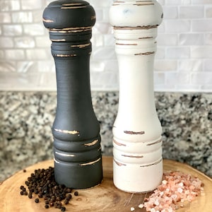 May include: A set of two salt and pepper shakers with distressed paint finishes. The shaker on the left is black with gold accents and the shaker on the right is white with gold accents. Both shakers have a classic design and are sitting on a wooden base.