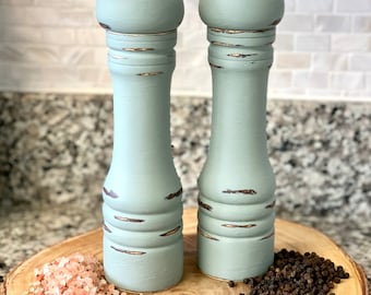 May include: Two matching, light blue, distressed wooden salt and pepper grinders with silver tops. They sit on a wooden slice with pink Himalayan salt and black peppercorns. The grinders are a kitchen accessory.