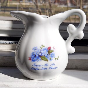 May include: White ceramic pitcher with a blue forget-me-not flower design. The pitcher has a handle and says "Forget-Me-Not Alaska State Flower".