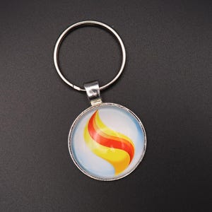 Puede incluir: Un llavero plateado con un colgante redondo. El colgante presenta un fondo blanco con un diseño en espiral en tonos rojos y amarillos. El llavero está sobre una superficie negra.