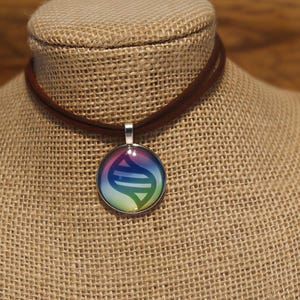 Puede incluir: Un collar gargantilla de cuero marrón con un colgante plateado. El colgante presenta un diseño colorido con tonos azules, morados, verdes y amarillos. El collar se exhibe en un maniquí cubierto de arpillera.