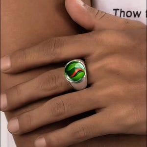 May include: A silver ring featuring a circular, domed cabochon inlay. The inlay is a vibrant, translucent green with a distinct, curved orange and black stripe pattern running through the center, resembling a classic glass marble design.