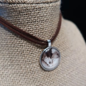 May include: A brown suede choker necklace with a silver pendant. The pendant features a black and white image of a cat. The necklace is displayed on a burlap surface.