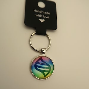 May include: A round, silver-toned keychain pendant with a colorful design featuring a rainbow gradient and a stylized symbol. The pendant is attached to a silver keyring and a black tag that reads "Handmade with love."