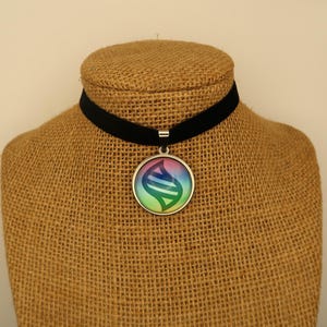 Puede incluir: Un collar gargantilla de terciopelo negro con un colgante plateado. El colgante presenta un diseño colorido y estilizado con tonos de azul, verde y rosa. El collar se exhibe en un maniquí beige.