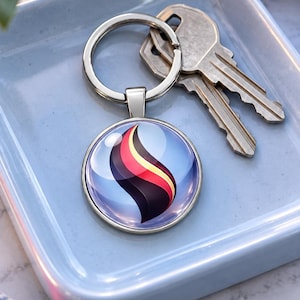 May include: A circular metal keychain featuring a glossy, domed glass cabochon with a stylized flame graphic in shades of black, red, and yellow against a light blue background, attached to a standard silver-toned split ring with two metal keys.