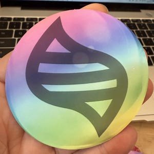 Puede incluir: Un botón redondo y colorido con un degradado de rosa, morado, azul, verde y amarillo. El botón presenta un emblema negro estilizado con forma de hoja y líneas geométricas, probablemente un artículo hecho por un fan.