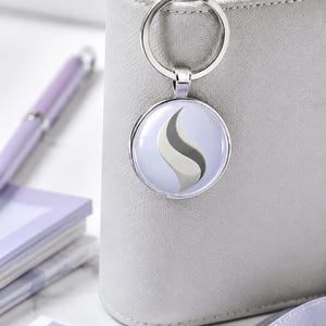 May include: A circular silver keychain featuring a light purple background with a stylized, curved gray and white flame-like graphic in the center. The keychain has a polished metal ring and a smooth, rounded frame that holds the printed design.