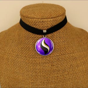 May include: A black velvet choker necklace featuring a round silver-toned pendant. The pendant displays a glossy purple circular design with a central abstract graphic element in black, white, and gold colors, suspended from a simple metal bail.