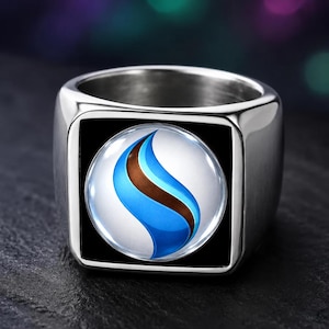 May include: A silver-toned metal ring featuring a square face with a circular inlay. The inlay displays a glossy white background with a prominent, stylized blue and brown flame-like graphic centered within the circle, set against a black square.