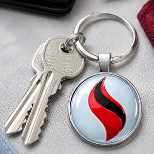 Może przedstawiać: Brelok do kluczy z metalu w kolorze srebrnym z okrągłą zawieszką o błyszczącym białym tle i stylizowanym czerwono-czarnym wzorem graficznym przypominającym płomień. Zawieszka jest przymocowana do standardowego kółka, na którym znajdują się dwa srebrne klucze.