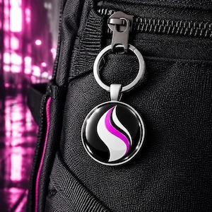 May include: A circular keychain with a silver metal frame attached to a black backpack zipper. The keychain features a glossy black background with a central graphic design consisting of a white curved shape and a vibrant pink curved stripe.