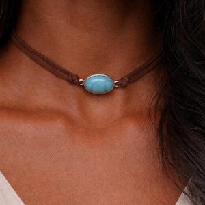 May include: A brown suede choker necklace with a turquoise oval stone pendant. The stone is set in a silver-colored metal. The necklace is worn around the neck.
